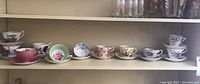 Eleven assorted vintage teacups and saucers arranged on shelves showing varied floral and scenic patterns with pastel colors and gold trim.