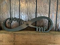 Front view of black metal infinity wall decor mounted against a dark wooden wall, showing the words 'you' and 'me' at either end of the infinity symbol.