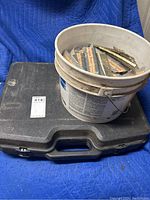 Closed black hard plastic carrying case with white plastic bucket of collated nails placed on top.