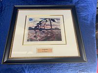 Framed artwork showing a colorful landscape with a tree bent by wind near water and hills, black frame with gold beading.