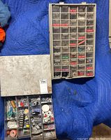 Two hardware organizers laid open on a blue surface. One organizer has a metal lid and is filled with various metal pins, connectors, springs, and small electronic parts. The other organizer has plastic compartments with red labels showing types of hardware including screws and plugs.