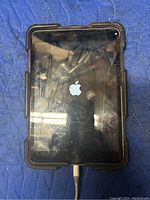 Front view of the Apple iPad with the Apple logo on screen and visible small crack, connected to charger.