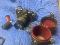 Asahi Pentax camera with attached zoom lens, two film canisters, a lens cap with red felt lining, and a carrying strap on a blue padded surface.