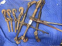 Photo showing two heavy-duty metal ratchet binder load binders with rust and wear, laid out on blue padded blanket with four separate heavy-duty spare hook ends with chain links.