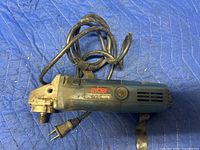 Ryobi 600W angle grinder placed on blue padded surface with visible power cord and two-prong plug. Angle grinder shows signs of use and has Ryobi branding on side.