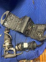 Electric angle grinder and reciprocating saw displayed on a blue moving blanket with cords wrapped around them, next to black toolbox containing hand tool accessories.