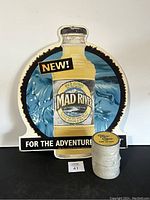 Cardboard advertisement shaped like a Mad River Wild Citrus bottle with 'NEW!' and 'FOR THE ADVENTURE' text, blue background with water imagery.