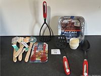 Cooking and baking utensils including spatulas, basting brush, potato masher, slotted spatula, disposable aluminum pans, and cupcake liners on a white surface