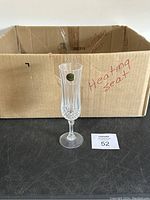 Single crystal champagne flute glass in front of cardboard box with 'heating seat' handwritten text, showing full view of glass including stem and base.