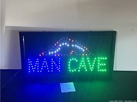 Illuminated LED sign spelling 'MAN CAVE' with multi-colored lights including blue, green and red, and a roof graphic above the text.