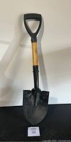 Mini shovel standing upright on black surface showing front side with wooden handle and black plastic D grip