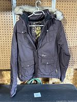 Front view of brown Orage jacket hanging on pegboard, showing flap pockets, zippered side pockets, and faux fur trimmed hood.