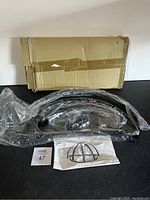 Photo of disassembled black metal curved pot rack components including hooks, mounting hardware, and instruction sheet, packaged in clear plastic with a cardboard box.