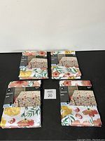 Full lot of four new Hometrends tablecloths with floral pattern on white background shown in packaging.