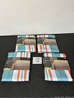 Four folded Hometrends tablecloths placed flat showing packaging with striped pattern in teal, orange, white, and beige colors.