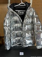 Front view of the silver Tna The Super Puff puffer jacket sized small hanging on a white hanger against a wooden pegboard background.