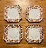 Four square cake plates with ornate floral and ornamental borders, gold rimmed center circles, placed on wooden surface.