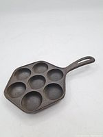Front top view of the pan, clearly showing 7 deep round wells and flat handle with slot for hanging. Dark seasoned cast iron finish visible.