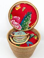 Overhead photo showing the vintage Chinese ceramic teapot with bamboo handle inside a round woven basket. The basket is lined with bright red fabric patterned with flowers, butterflies, and peacock feathers. The teapot lid is placed near the handle, and a ceramic spout piece is next to the teapot inside the basket.