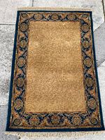 Full view of rectangular wool rug with beige center and floral dark blue border with fringe ends laid flat on concrete.