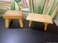 Two rustic wooden stools shown together on black surface with painted green backdrop. One has thick legs and block style while the other has three legs and a flat top.