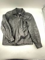 Front view of black leather jacket laid flat showing zipper closure, collar, side pockets, and sleeves with snap button cuffs.