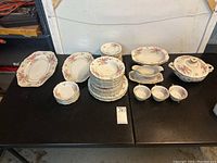 Photo showing full partial china set arranged on black table, including stacks of plates, small bowls, oval serving platter, gravy boat, small covered dish, and other serving pieces.