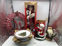 Photo showing entire Christmas decoration lot including doll in box, wreath with candles, stacked Christmas plates, and two Santa figurines.