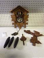 Full view of cuckoo clock body, detached pine cone-shaped weights, pendulum with leaf piece, carved wooden bird, and additional carved decorative wooden pieces laid out on flat surface.