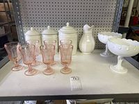 Overview of entire lot including three white ceramic cooking jars with lids, six pink pressed glass goblets, white ceramic pitcher, and two white milk glass pedestal bowls on a white table.