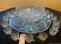 Picture showing the cut glass punch bowl centered on an 18 inch round drip platter, surrounded by 18 cut glass cups evenly spaced around the perimeter on a dark surface.