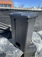 Photo of the Safco black plastic indoor trash can, showing rectangular shape with a closed lid and step pedal at bottom front.