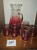Photo showing vintage cranberry glass pitcher with six matching glasses on wooden surface, with handwritten number card.