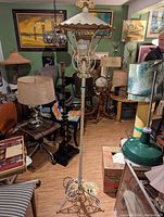 Full view of 1950s patio floor lamp with decorative metal scrollwork and scalloped canopy shade, displayed standing on a floor surrounded by other vintage furniture and lamps.