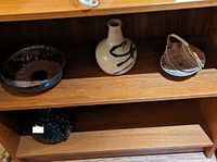 Three ceramic items on a wooden shelf including a dark bowl on the left, a beige vase in the middle, and a light brown container with a handle on the right.
