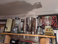 Six vintage kitchen appliances displayed on a wooden shelf, including various blenders, coffee makers, and a milkshake maker, with a framed artwork on the wall behind them.