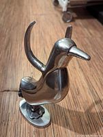 Photo showing a shiny aluminum penguin figurine with one wing raised, standing on an oval base on a wooden floor.