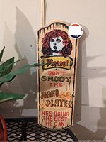 Photo of a wooden bar sign with a vintage woman illustration and text "Please! Don't Shoot The Piano Player He's Doing The Best He Can."