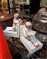 Photo showing two matching ceramic child figurine bookends on a black wire shelf with various objects around.