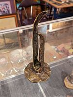 Photo of a 6 inch tall bronze figure with an abstract, elongated form mounted on an irregular shaped base, displayed on a glass counter with other antiques in background.