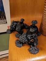 Two black ceramic poodles with detailed textured fur on ears, head, legs, and tail, positioned lying down on wooden surface.