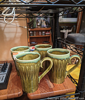 Photo of four ceramic mugs grouped together on a wooden table under a black metal shelf, showing textured green exterior and light blue glaze inside.
