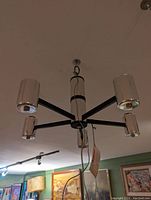 Photo shows chrome chandelier with five cylindrical shades mounted on black metal arms hanging from ceiling in room with artwork on walls.