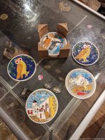 Top-down view of 6 colorful round coasters with various hand-painted style designs, including owls and scenic Mediterranean village scenes, arranged on a glass surface with the wooden holder in the center.