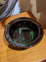 Photo showing a round ceramic dish with black base and green and brown glaze drips on a wooden surface.
