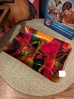 Photo showing a small rectangular glass dish with an abstract, colorful enamel design placed on a fabric surface, with a vintage science-themed book partly visible in the background.