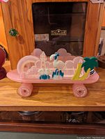 Pink skateboard-shaped cassette tape holder featuring decorative flamingo, palm tree, rainbow, and lighthouse on top, photographed on wooden surface with background cabinet