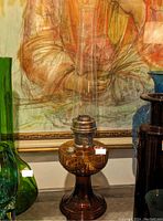 Brown glass oil lamp with clear glass chimney and metal wick holder standing on a surface against a background with framed artwork and other glass items.