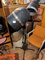 Photo showing two vintage hair dryer heads mounted on adjustable metal stands with tripod bases, converted into lamp fixtures in a room setting with orange chair and other furniture.