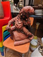 Heavy cement garden gnome statue placed on an orange chair, showing detailed face, beard, hat, and seated pose on a log base.